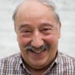 Elderly man with mustache smiling at the camera in an urban outdoor setting.