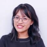 Portrait of a young woman with glasses and black hair smiling against a plain background.