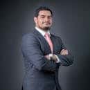 Professional portrait of a confident businessman in a gray suit with a pink tie, posing with arms crossed.