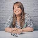 Attractive female with long hair smiling and holding pen while sitting at table with glasses and looking away on white background