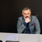 A thoughtful businessman in a suit working on his laptop in a modern office setting.